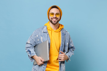 Canvas Print - Smiling young hipster guy in fashion jeans denim clothes posing isolated on pastel blue background studio portrait. People emotions lifestyle concept. Mock up copy space. Holding laptop pc computer.