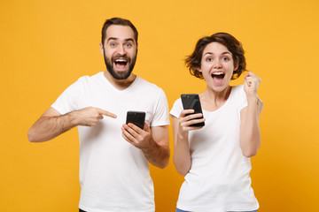 Excited couple friends bearded guy girl in white t-shirts isolated on yellow orange background. People lifestyle concept. Mock up copy space. Point index fingers on mobile phone, doing winner gesture.