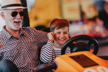 Grandfather And Grandson Having Fun And Spending Good Quality Time Together In Amusement Park. They Enjoying And Smiling While Driving Bumper Car Together.