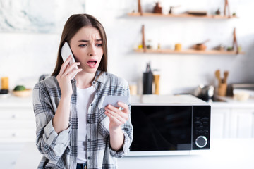 Wall Mural - shocked woman talking on smartphone near microwave and holding Business Card in kitchen