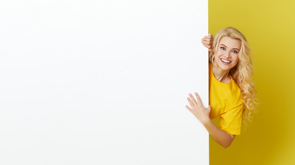 Wall Mural - Young happy woman peeks out from behind a white banner on a yellow background. Point to an empty blank on a form, a copy space for text. Horizontal shot