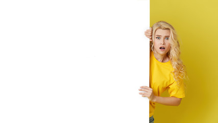 Wall Mural - Young happy woman peeks out from behind a white banner on a yellow background. Point to an empty blank on a form, a copy space for text. Horizontal shot