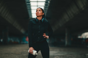 Young woman in black sportswear under overpasses resting after jogging in cold weather holding protein shake