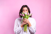 Studio Portrait Of Gorgeous Young Brunette Woman With Long Wavy Hair Wearing White Loose Cotton Shirt, Holding Bouquet Of Tulip Flowers. Pink Isolated Background, Copy Space, Close Up.