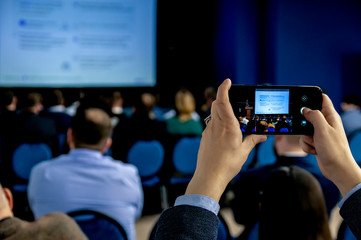 Men's hands in business suit holding smartphone and taking pictures of the screen with business presentation at official conference, meeting in crowded room.