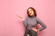 Positive lady with curly red hair and gray sweater isolated on a pink background, holds a blank space in her hand and looks in camera with a smile on her face.Happy girl holding copy space on her hand