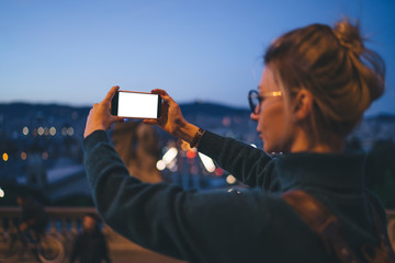 Back view of millennial hipster girl in casual apparel taking pictures of evening city via smartphone application with copy space touchscreen, female tourist photographing urban setting via cellphone