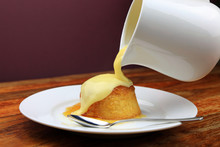 Hot custard being poured from a jug over a treacle sponge pudding.