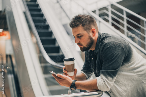 Cuadros en Lienzo Young handsome man in shirt walking in city mall with cup of coffee and smartphone