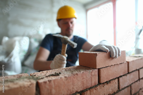 Fotomural Portrait of skilled man constructing big concrete wall and using special hammer to properly fulfill task to gently lay bricks on unfinished structure