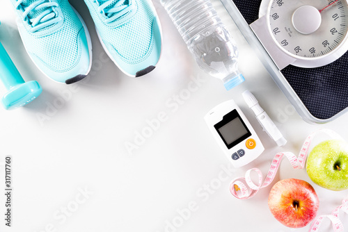 Healthy lifestyle, food and sport concept. Top view of diabetes tester set with athlete\'s equipment Weight Scale measuring tape green dumbbell, sport water bottles, fruit on white background.