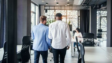 Serious caucasian and afro american businessmen coming in the modern office room and discussing details of their todays job.