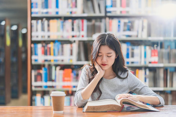 Wall Mural - Asian young Student in casual suit reading the book with a cup of coffee in library of university or colleage on the wooden table over the book shelf background, Back to school concept