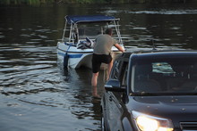 White Fishing Motor Boat Launch, A Caucasian Man Puts A Boat On The Black Car Trailer In Water On Slipway