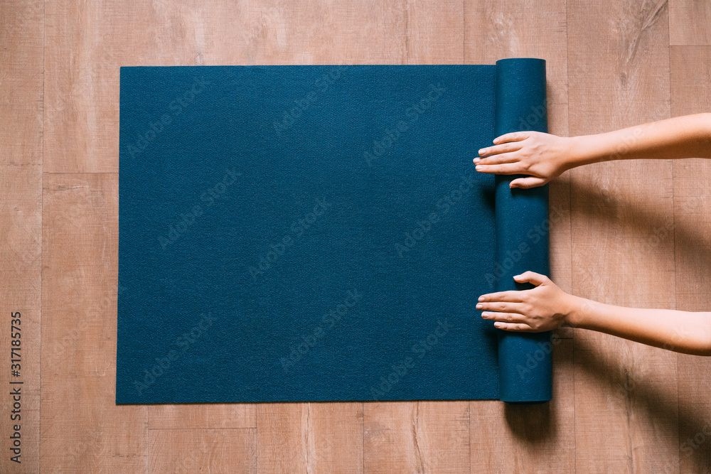 Fit woman folding blue exercise mat on wooden floor before or after working out in yoga studio or at home. Equipment for fitness, pilates or yoga, well being concept. Flat lay, space for text. - obrazy, fototapety, plakaty 