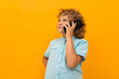 Little boy with curly hair in blue shirt and shorts calls the phone isolated on yellow background
