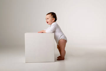 Wall Mural - baby boy in white bodysuit stands leaning on a white cube on a light background, space for text