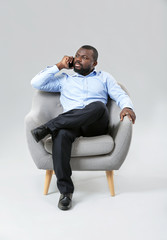 Poster - African-American man talking by phone while sitting in armchair against grey background