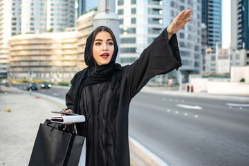 Wall Mural - Beautiful arab woman in Abaya with shopping bags, phone and wallet hitching a taxi on the street