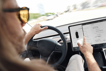 Mode of Transport. Bearded man with long hair in sunglasses traveling sitting inside electric car driving choosing settings on smart system concentrated close-up