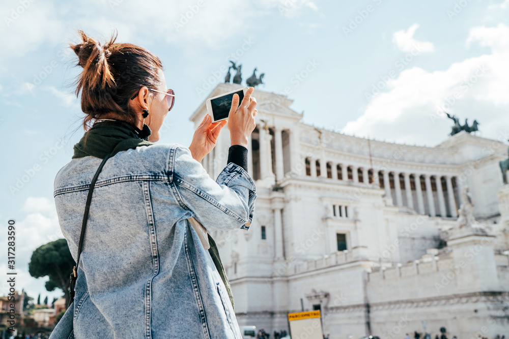 Young alone caucasian female tourist taking a photo of Victor Emmanuel II Monument using a modern smartphone on Piazza Venezia in Rome, Italy. - obrazy, fototapety, plakaty 