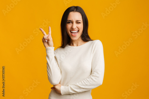 Fotografía Playful brunette girl showing peace sign and winking on yellow background