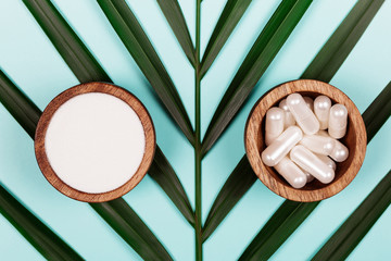 Wall Mural - Collagen in capsules and in powder in two wooden bowls on tropical leaf background