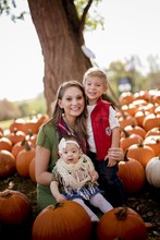 Woman With Two Little Kids Sitting In A Garden With Pumpkins On The Blurry Background