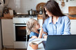 Working mom works from home office. Happy mother and daughter look to each other. Woman and cute child using laptop. Freelancer workplace in cozy kitchen. Female business, kindness, care. Lifestyle.
