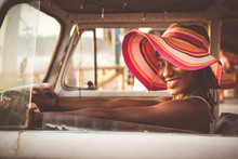 Side View Of Cheerful Young African American Female In Colorful Wide Brimmed Striped Hat Sitting Behind Wheel Of Vintage Van And Looking At Camera