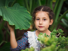 Little Girl In Retro Dress Trying Piece Of Fruit While Standing Near Green Bushes On Summer Day In Garden