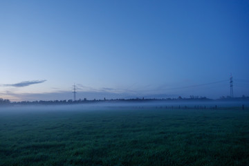 Beautiful dramatic twilight gold and blue cloud and sky over misty atmosphere of foggy over agricultural field and high voltage tower on countryside in Germany during sunset time.