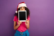 Positive Mixed Race Girl In Santa Hat Looking At Camera While Showing Digital Tablet With Blank Screen On Purple Background