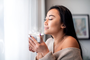 Wall Mural - Relaxed asian woman with cup of tea or coffee spending leisure time at home