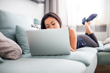 Wall Mural - Smiling asian young woman using laptop lying on sofa at home