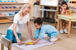 © LIGHTFIELD STUDIOS - Selective focus of teacher with kid playing on floor and boring child at desk in montessori school