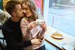 © vitaliymateha - Happy romantic couple sitting over the window in cafe, embracing, kissing and drinking coffee. boyfriend tenderly hugging girlfriend in cozy cafe.