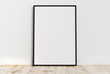 © Oleksandr - Blank vertical black poster frame standing on light wooden floor with next to white wall. Blank poster frame mockup. Empty picture frame mockup. Vertical frame mock up. Blank photo frame. 3d rendering