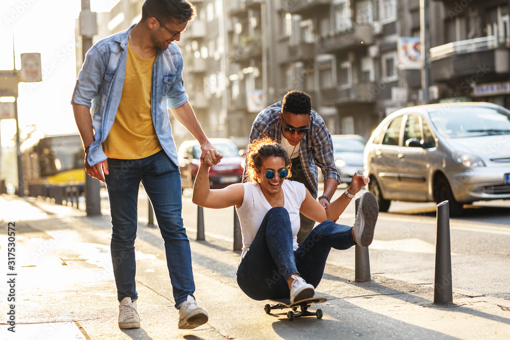 Group of friends hangout at the city street.Female sitting on skate board while friends pushing her from behind.	 - obrazy, fototapety, plakaty 