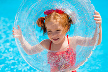 Little Red-haired Girl In A Red Swimsuit And Heart - Shaped Sunglasses Holds A Swimming Circle In The Pool