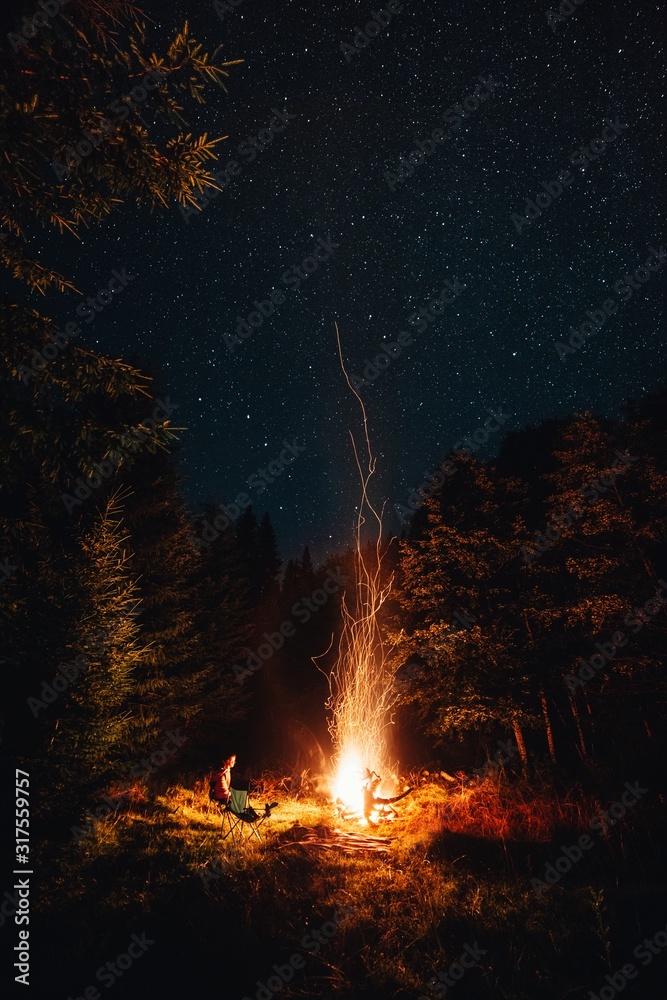 Young female sitting on chair in the wilderness forest near the big campfire under the milkyway.  - obrazy, fototapety, plakaty 