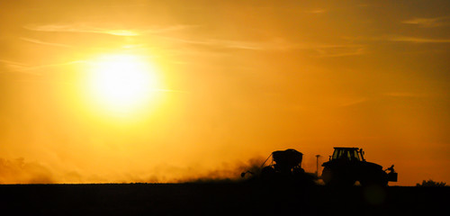 Silhouette of a tractor sowing seeds in a field in a cloud of dust against the background of the setting sun.