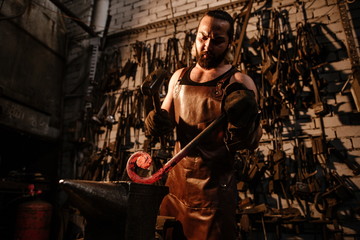 Brutal muscular artisan blacksmith standing in the workshop with a hammer, banging on a hot rod of iron on an anvil