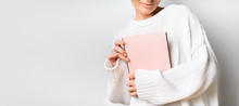 Close View Of Woman In White Woolen Sweater Holding A Book With Empty Pink Cover In Hands. Free Space For Your Mock Up Of Reading Book Concept Background.