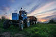 Old blue tracktor with a trailer on green grass. Evening, sunset. Beautiful colorful sky.