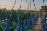 Fototapeta Na sufit - Hanging (suspension) bridge across a narrow river. Blue iron fences and old wooden bottom. Summer. Green trees and meadows. Clear blue sky. Evening. Middle Russia. Kaluga region
