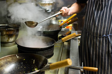 Chefs At Work In A Commercial Kitchen. Male Chef Cooking Using Wok Styled Pans In A Takeaway / Restaurant Setting
