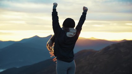 back view of hipster millennial young woman on top of mountain summit at sunset raises arms into air