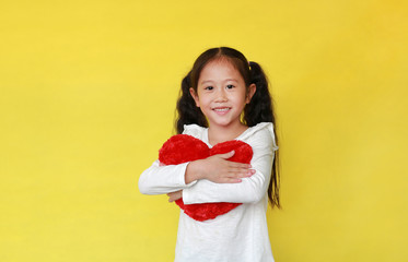 Wall Mural - Portrait of happy asian child girl hugging a fluffy red heart with looking at camera isolated on yellow background.