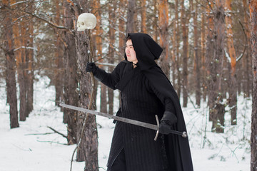 Warrior, magician in black quilted clothes and in a cloak with a hood against the backdrop of a winter forest holds a sword and a staff with a skull in his hands.